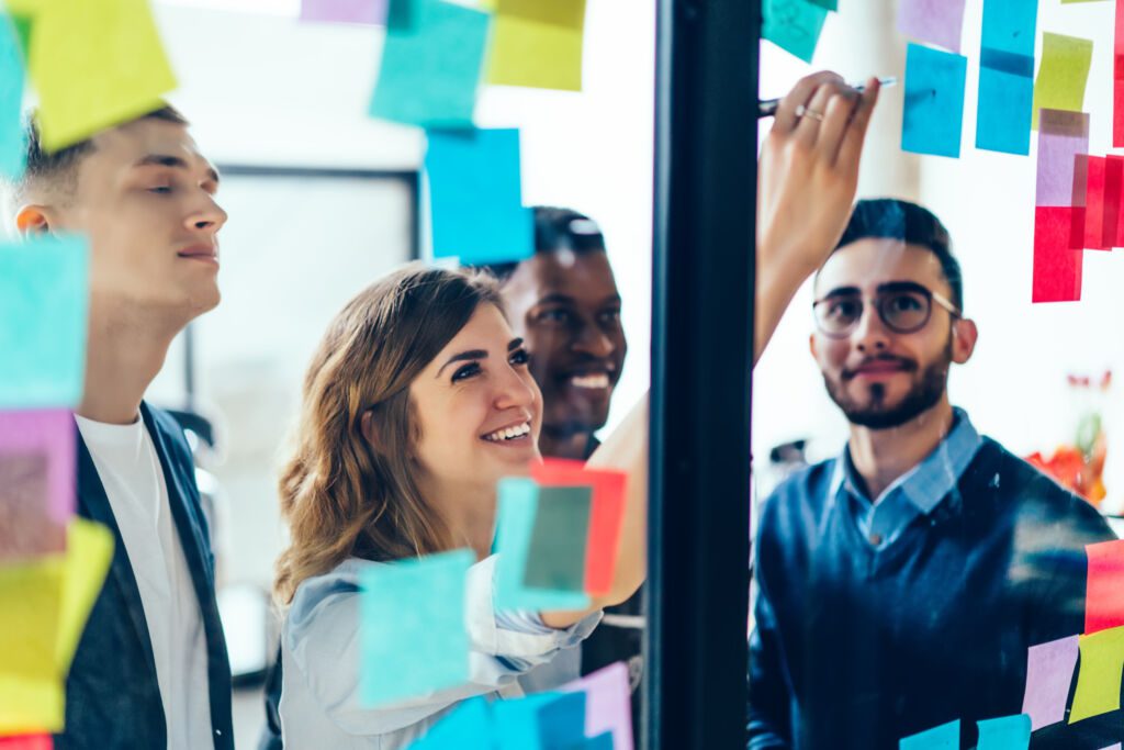 Diverse team of positive young people laughing while working together during brainstorming and standing behind glass wall with sticky colorful papers.