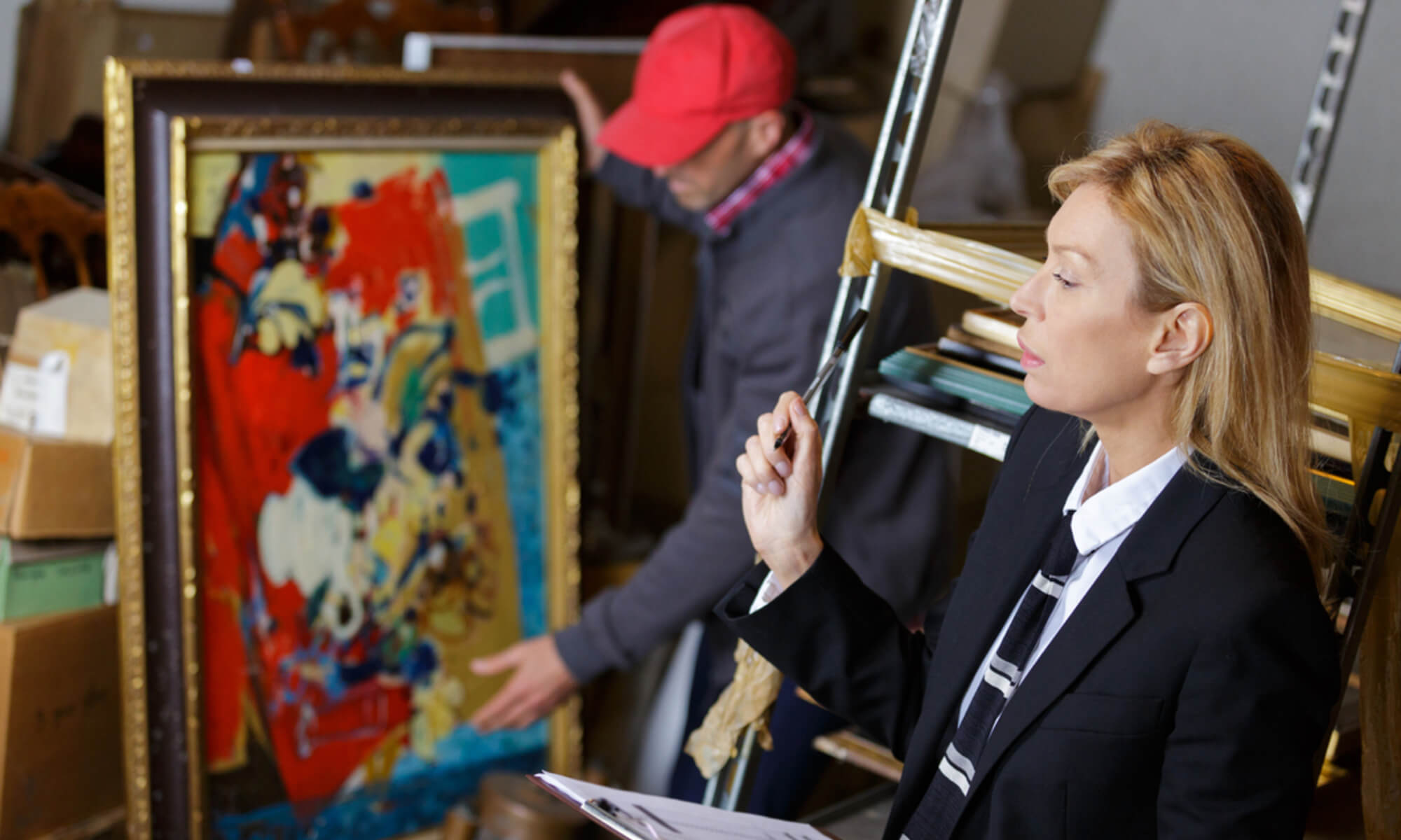 A woman with a bid sheet in the foreground and a man showing off a painting in the background.